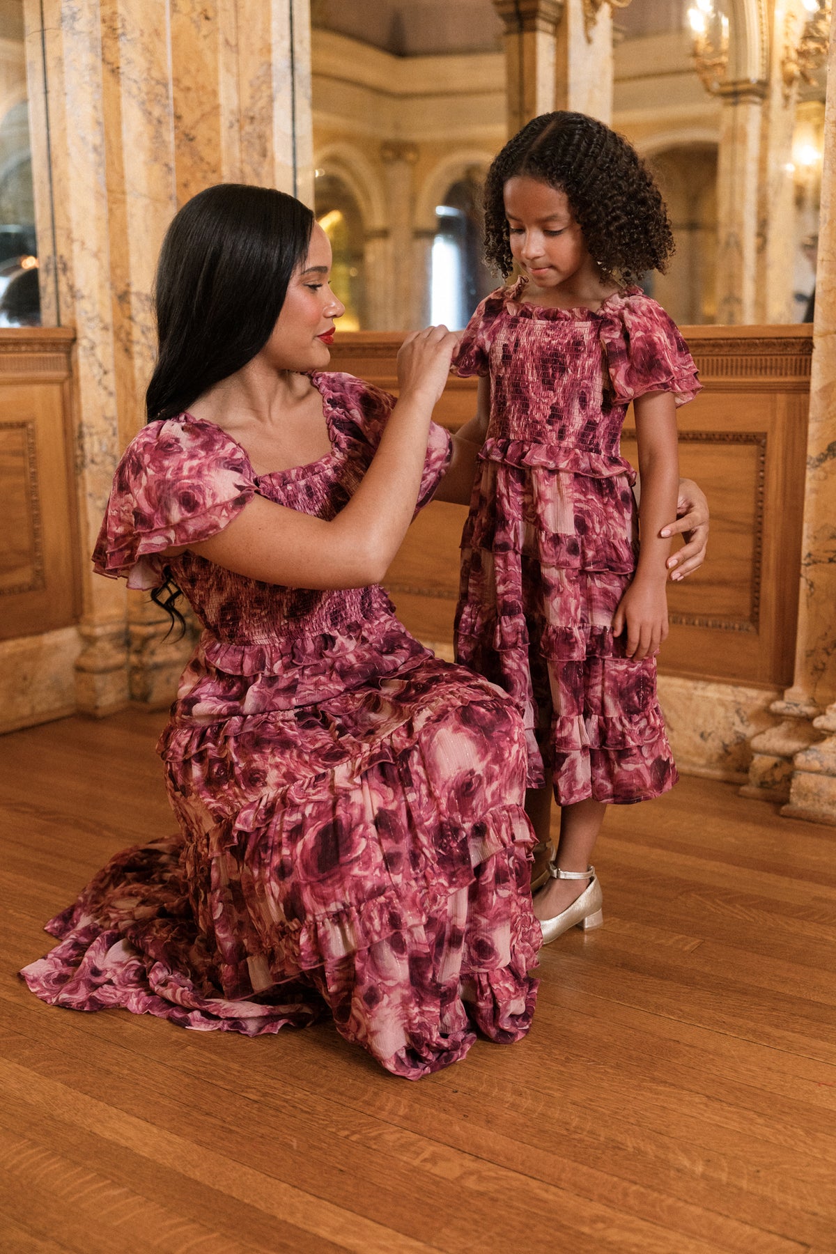 A woman and a young girl, both dressed in Ivy City Co’s Winnie Dress in Dreamy Roses, stand on a wooden floor in an elegant room adorned with marble columns and ornate decor. The woman kneels, smiling as she adjusts the girl’s holiday dress, ensuring the gold metallic stripe on their matching chiffon outfits shimmers beautifully in the light.