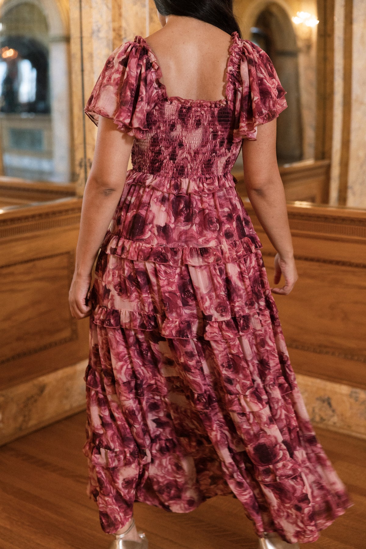 A woman stands indoors with her back to the camera, enveloped in the Ivy City Co Winnie Dress in Dreamy Roses—a flowing pink floral chiffon dress detailed with delicate gold metallic stripes—surrounded by ornate mirrors and rich wood paneling, evoking the essence of a dreamy holiday or elegant girls’ dress inspiration.