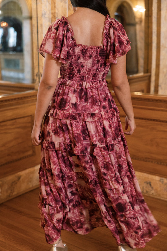 A woman stands indoors with her back to the camera, enveloped in the Ivy City Co Winnie Dress in Dreamy Roses—a flowing pink floral chiffon dress detailed with delicate gold metallic stripes—surrounded by ornate mirrors and rich wood paneling, evoking the essence of a dreamy holiday or elegant girls’ dress inspiration.