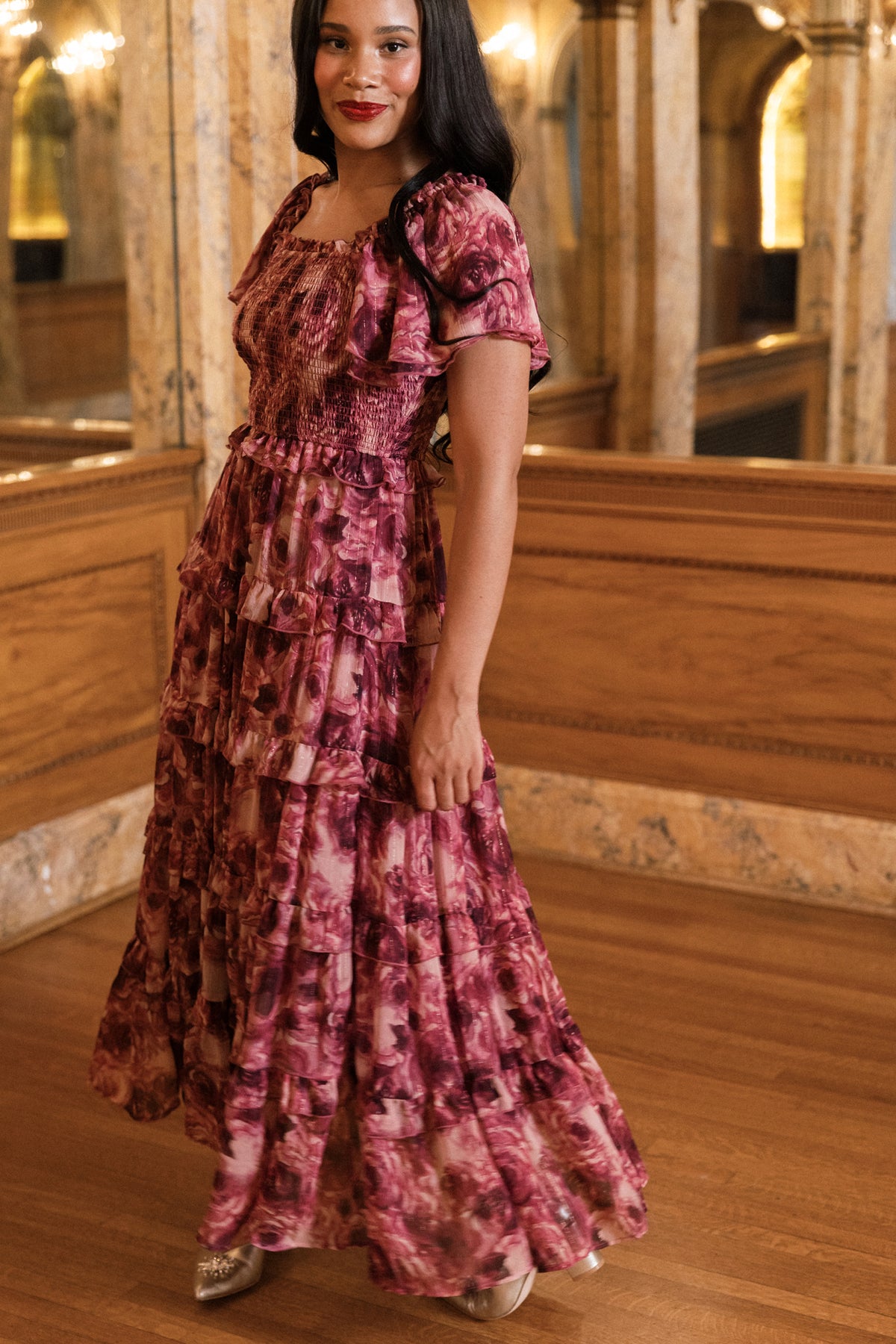 Wearing the Winnie Dress in Dreamy Roses by Ivy City Co, a woman stands indoors on a wooden floor, surrounded by ornate mirrors and columns. She smiles gently at the camera as subtle gold metallic stripes on her flowing pink floral holiday dress catch the light.