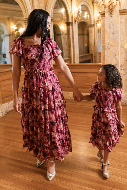 An adult and a child, both dressed in the Winnie Dress in Dreamy Roses by Ivy City Co—with matching floral chiffon fabric and a subtle gold metallic stripe—hold hands as they walk together through an elegant, ornate building adorned with marble columns and wooden floors, creating the perfect scene for their stunning holiday dress ensemble.