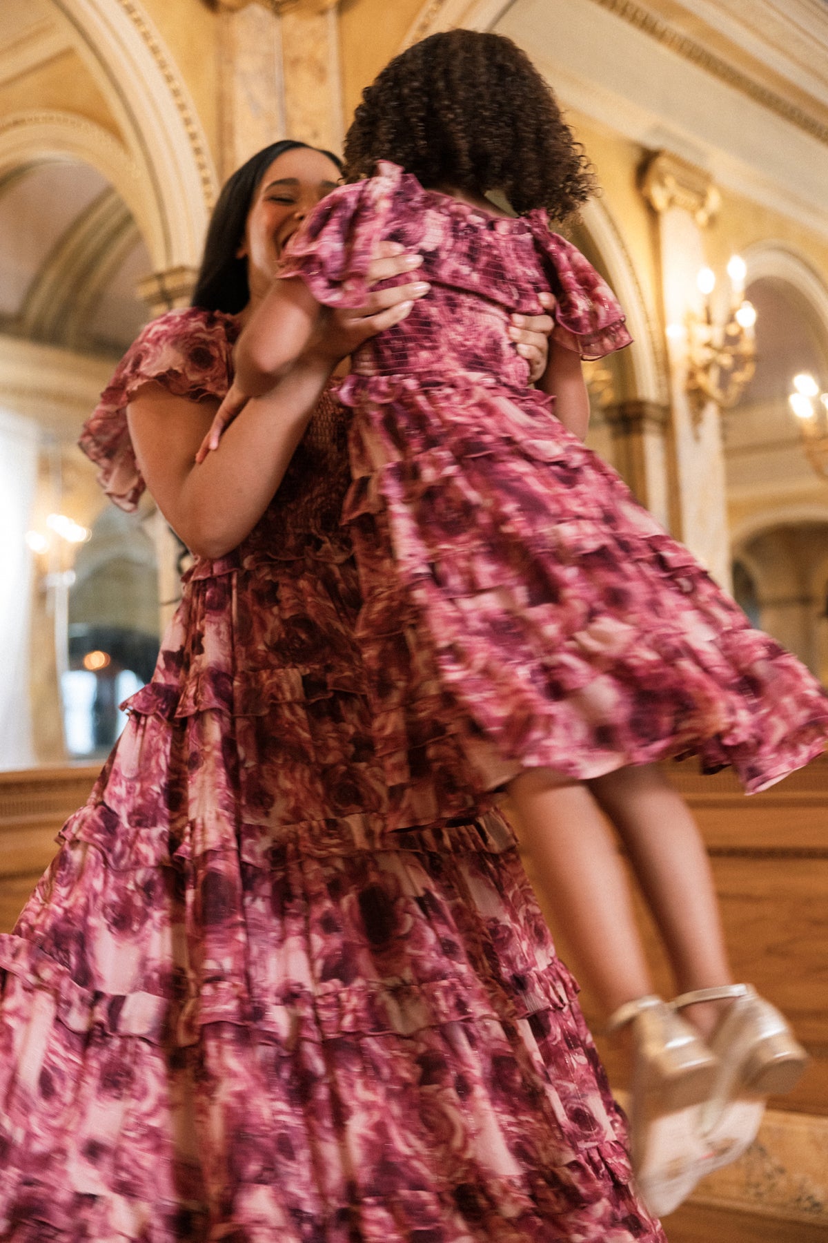 A woman lifts a child in the air; both wear matching Ivy City Co “Winnie Dress in Dreamy Roses” dresses—pink florals with gold metallic stripes—in an elegant, ornate room featuring arched ceilings and gold accents.