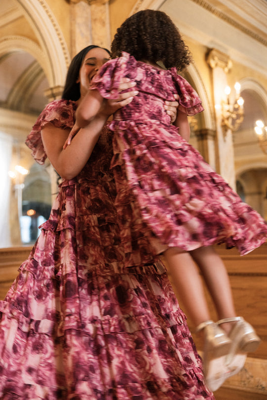A woman lifts a child in the air; both wear matching Ivy City Co “Winnie Dress in Dreamy Roses” dresses—pink florals with gold metallic stripes—in an elegant, ornate room featuring arched ceilings and gold accents.