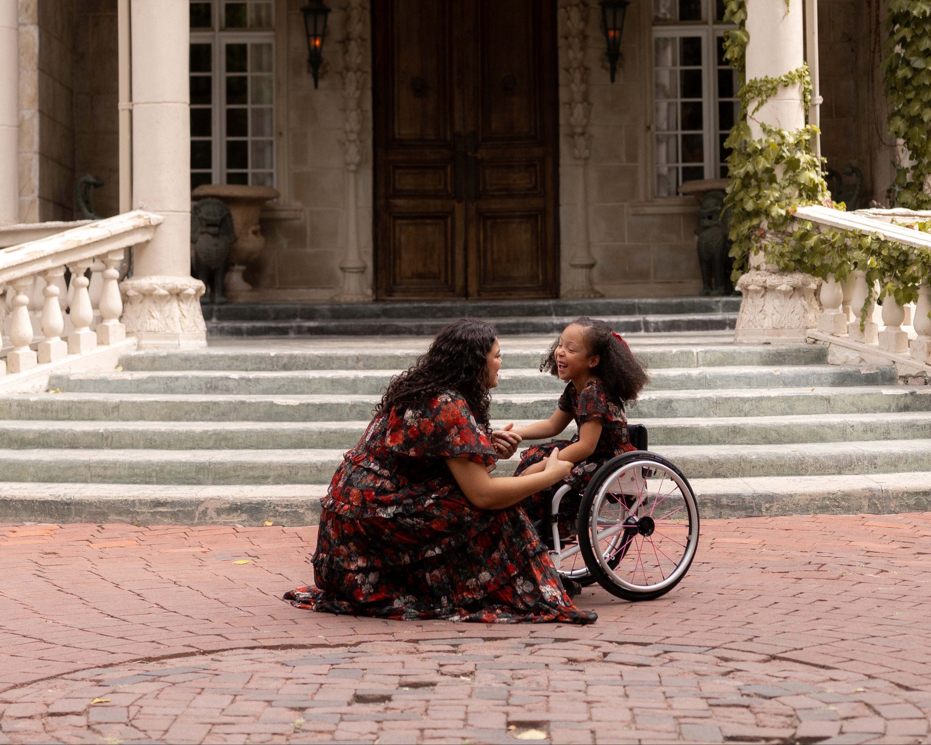 Woman sitting on a stone path with a child in a wheelchair in front of a large building.