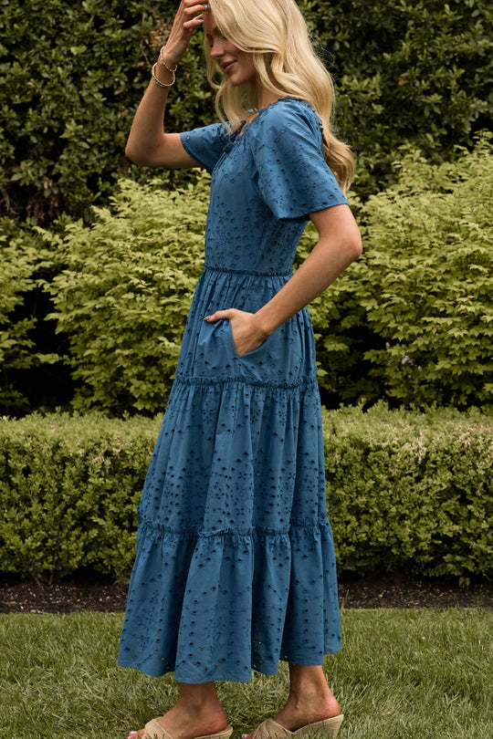 A woman with long blonde hair wears the Ivy City Co Etta Dress in Cobalt—a short-sleeve, ankle-length dress with eyelet details—while standing outdoors in front of green bushes.