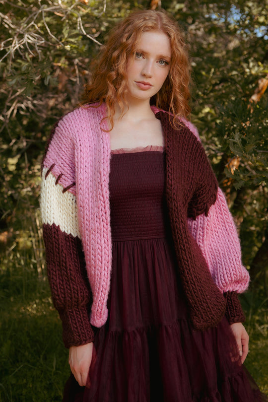 A woman with long red hair wears the Ivy City Co Marissa Sweater in Pink & Red layered over a dark strapless dress, standing outdoors among green trees and sunlight.