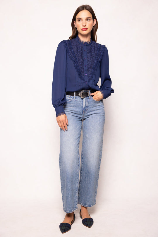A woman with straight brown hair and a neutral expression stands against a white background, wearing the Ivy City Co Blythe Blouse in Navy—featuring nursing-friendly design and lace details—paired with light blue wide-leg jeans, a black belt, and pointed-toe flats.