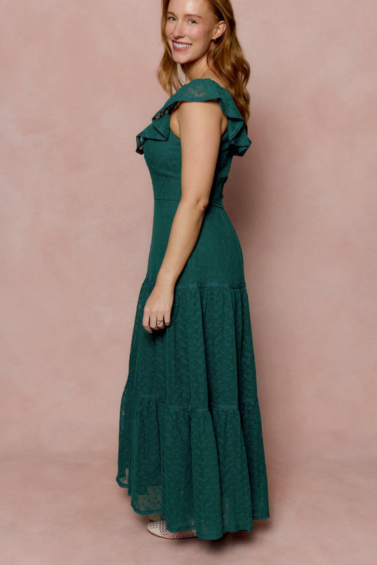 A woman stands sideways and smiles in front of a soft pink background, wearing the Ivy City Co Alaina Dress in Deep Sea Lace—a sleeveless, ankle-length dress with ruffle detailing.