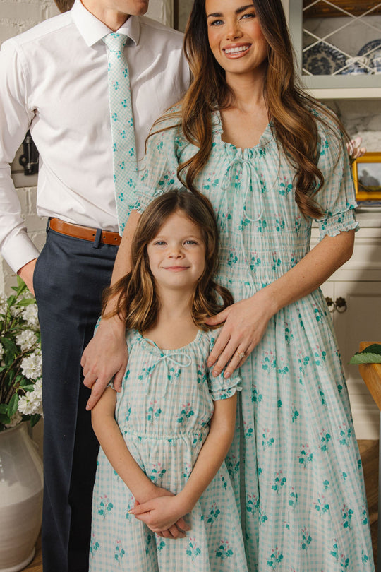 A man, woman, and young girl stand together indoors, each dressed in coordinated white and mint green floral outfits. While the woman and girl are smiling and the man’s face is partially out of frame, the young girl’s adorable look is accentuated by her Ivy City Co Mini Jessie Dress in Bloom Gingham—a spring dress featuring charming details perfect for the season.