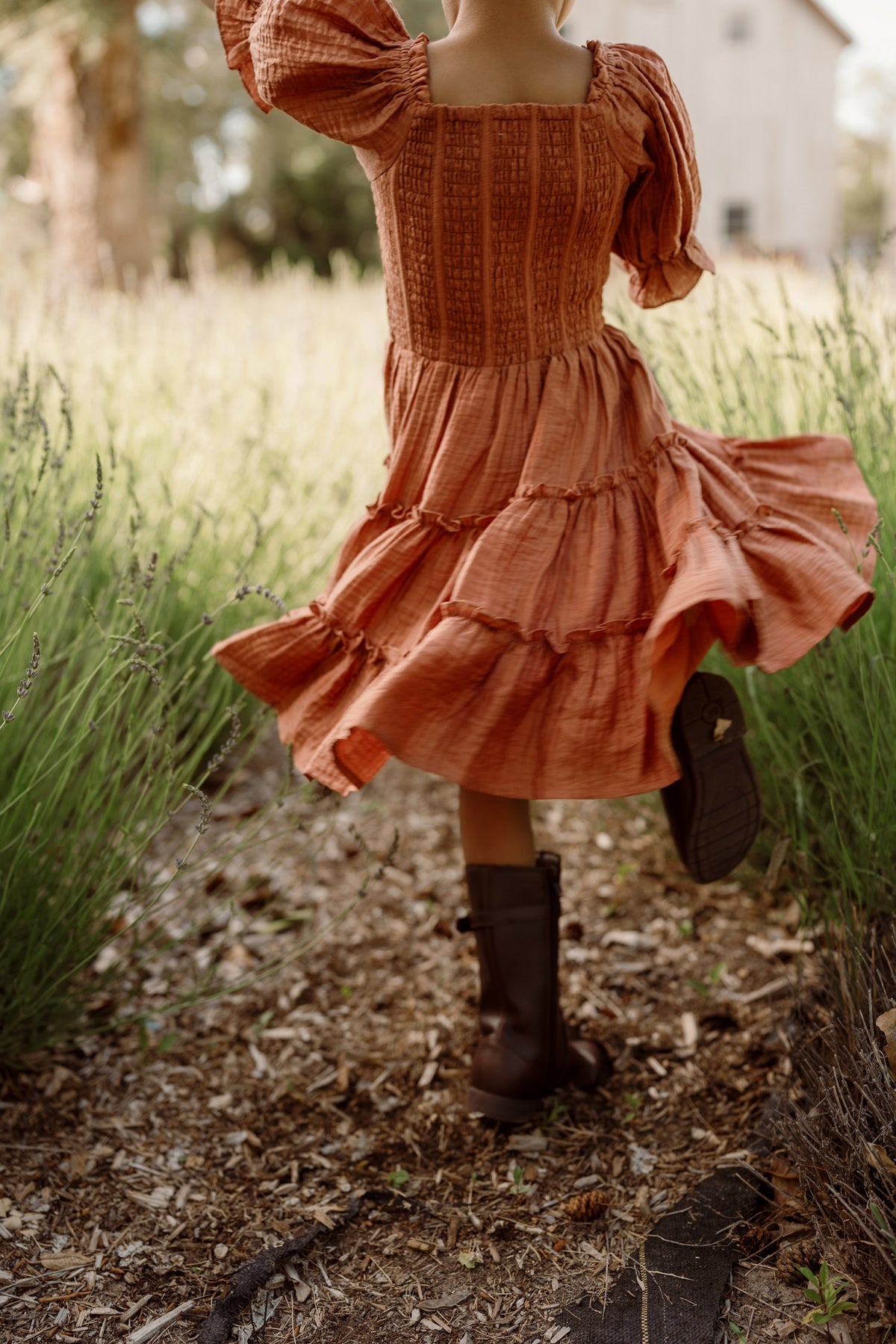 A child in a flowing Mini Madeline Dress in Apple Cider by Ivy City Co, featuring a smocked bodice and puff sleeves, twirls joyfully on a sunlit path through tall green grass in the countryside, her playful look finished with brown boots.