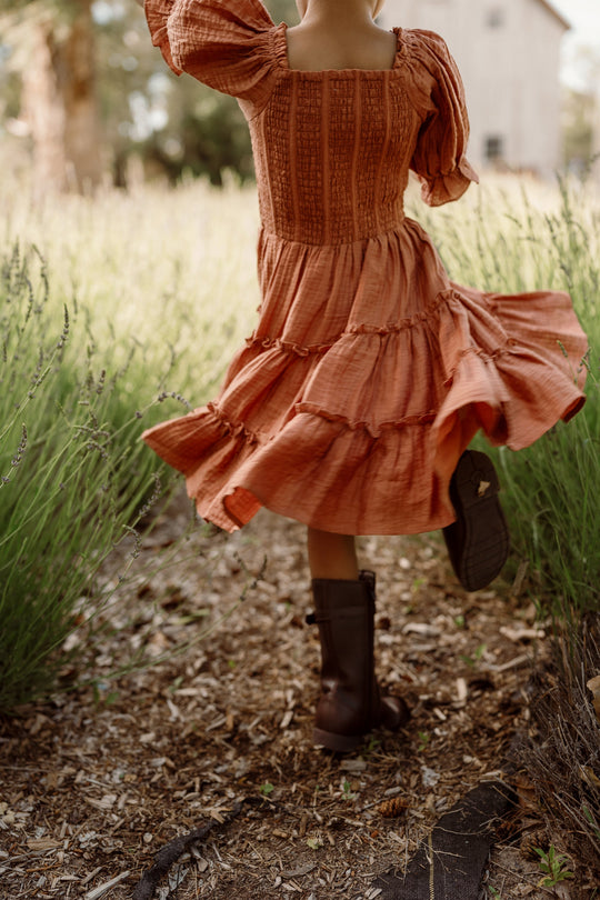 A child in a flowing Mini Madeline Dress in Apple Cider by Ivy City Co, featuring a smocked bodice and puff sleeves, twirls joyfully on a sunlit path through tall green grass in the countryside, her playful look finished with brown boots.