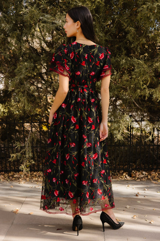 A woman stands outdoors with her back turned, wearing the Ivy City Co Flora Dress in Romantic Roses—a sophisticated black dress adorned with embroidered red roses and a delicate black mesh overlay. She finishes the look with black high heels, standing on a sidewalk bordered by greenery and framed by a black fence in the background.