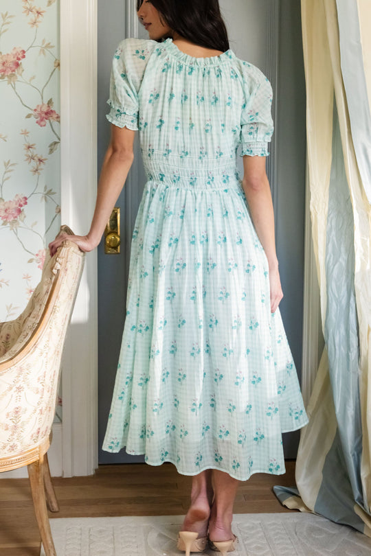 A woman stands indoors with her back to the camera, wearing the Jessie Dress in Bloom Gingham by Ivy City Co—an ankle-length, light blue dress featuring a charming floral pattern and short puffed sleeves. This maternity-friendly style combines comfort and elegance as she rests one hand on an upholstered chair near a doorway.