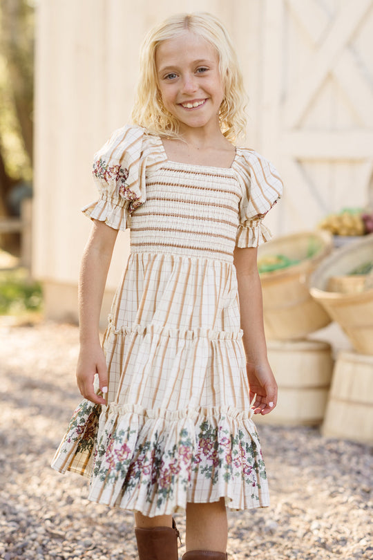 A blonde girl smiles outdoors, wearing the Ivy City Co Mini Madeline Dress in Embroidered Cider Grid, featuring a smocked bodice and puff sleeves. Wooden baskets filled with produce are visible in the background.