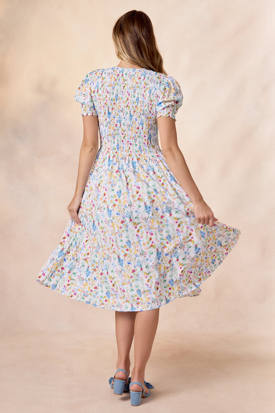 A woman stands with her back to the camera, holding out the edge of her Amelie Dress in Floral Fields by Ivy City Co against a neutral background. Her blue heeled shoes beautifully complement this nursing-friendly midi dress, which is designed for both style and comfort.