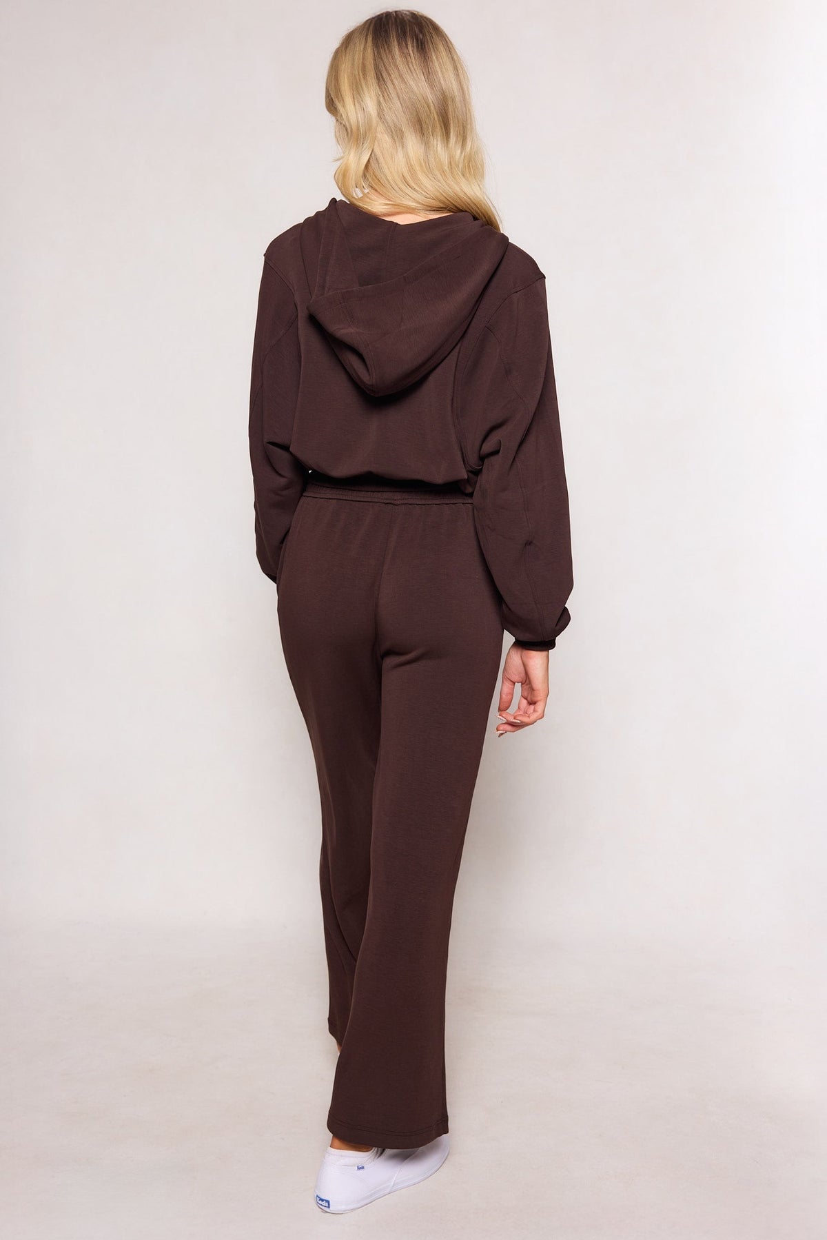 A person with long blonde hair, dressed in a dark brown hooded sweatshirt and the Ivy City Co Amara Sweatpant in Espresso—which features an elastic waistband and relaxed leg—stands facing away from the camera against a plain light background.
