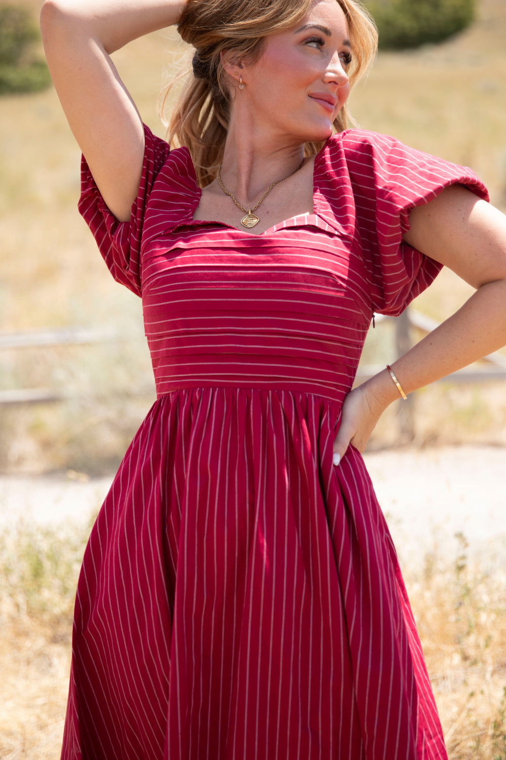 A woman wearing the Ivy City Co Julie Dress in Cranberry Pinstripe—with puff sleeves and a smocked bodice—stands outdoors in a sunlit grassy area, gazing to the side with one hand behind her head and the other on her hip, against a blurred backdrop of trees.