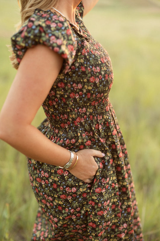 A woman stands outdoors in a field, her hand tucked into the pocket of the Ivy City Co Annalise Dress in Obsidian Vintage Poppies. The short-sleeved, floral dress—both elegant and maternity-friendly—is complemented by her bracelet and necklace, while her face remains out of frame. The background is a blur of green grass.