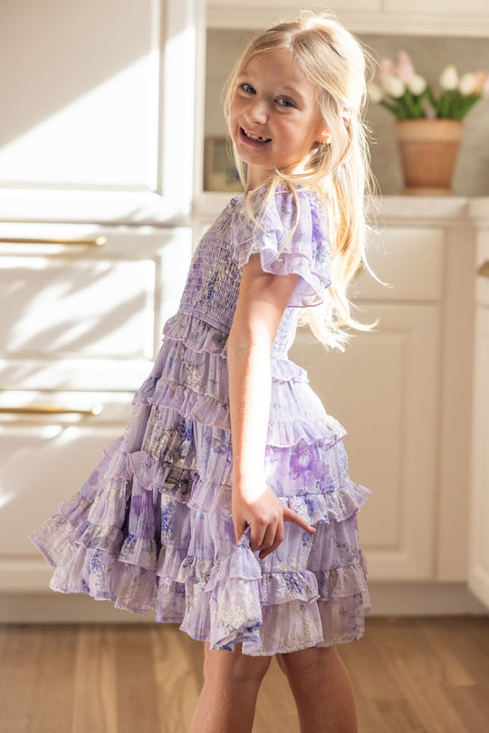 A young girl with long blonde hair smiles while posing indoors, sunlight illuminating her in a bright kitchen with white cabinets and a potted plant in the background. She wears the Ivy City Co Mini Winnie Dress in Lavender Aquarelle Fleur, featuring a smocked bodice and a delicate ruffle tiered skirt that adds charming detail to the scene.
