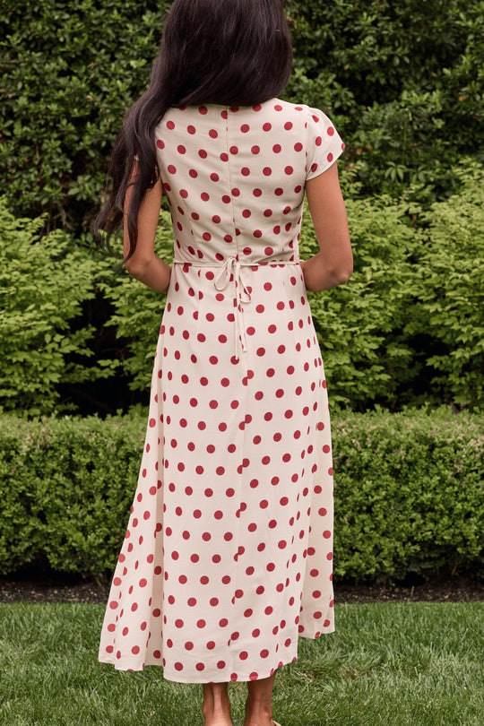 A woman with long dark hair stands outdoors, her back to the camera, wearing the Ivy City Co Fiona Dress in Red Polka Dot—a charming cream-colored, short-sleeve summer midi dress with a tie-back detail. Lush green bushes and grass surround her.