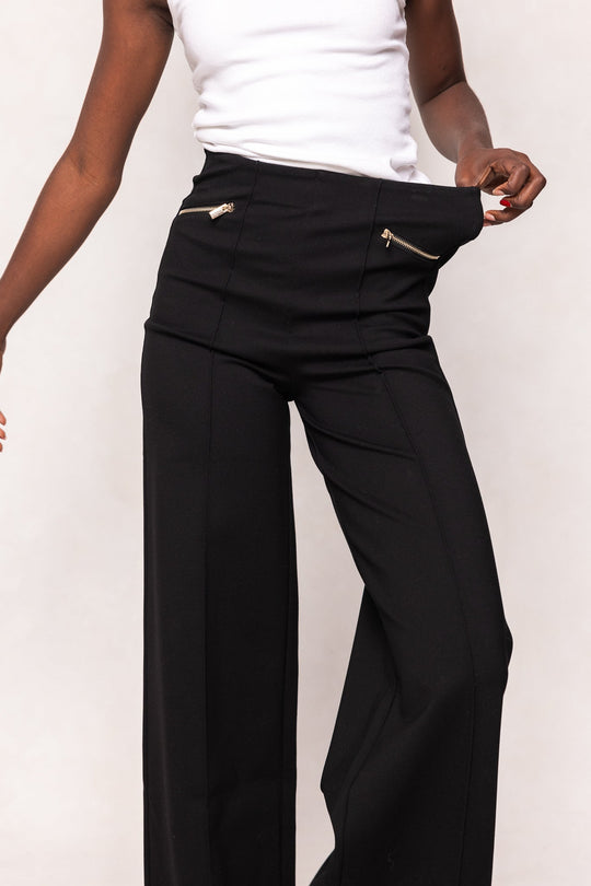 Wearing a white tank top, a person showcases the Ivy City Co Alyssa Pop Ponte Pants in Black, highlighting the gold zipper pockets and demonstrating the comfortable tummy control by pulling at the waistband to reveal extra space—making these loose-fitting pants an essential day-to-night staple.