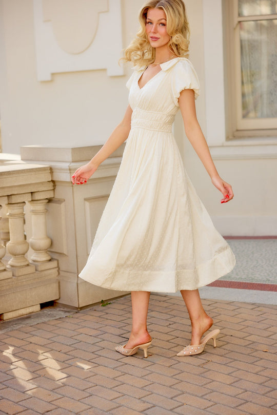Wearing the Zella Bow Dress in Ivory by Ivy City Co—a vintage-inspired, short-sleeve midi dress—paired with beige heels, a woman strolls outdoors on a brick path beside a light-colored building, her soft smile and elegant ensemble making it an ideal choice for a rehearsal dinner.