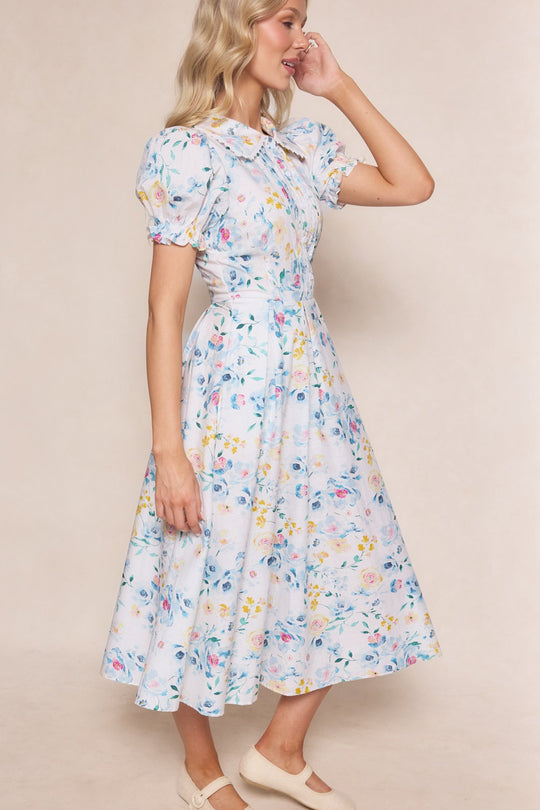 A woman with blonde wavy hair, standing sideways and touching her face, models the Sophia Dress in Blue Bouquet by Ivy City Co—a nursing-friendly midi dress featuring puff sleeves and a bow at the neck. She pairs it with cream-colored shoes, showcasing a look that is both stylish and practical.