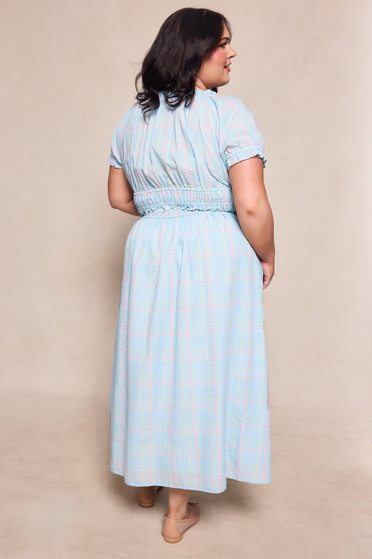 A woman with dark hair stands facing away from the camera against a beige background, wearing the Ivy City Co Thalia Dress in Pastel Plaid—a light blue, short-sleeved spring dress that pairs perfectly with her nude flats.