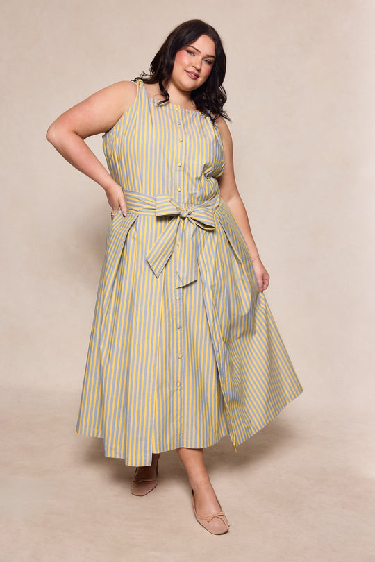 A woman wearing the Ivy City Co Glen Dress in Green Stripe—a sleeveless, ankle-length dress featuring green and white stripes with a large bow at the waist—stands confidently with one hand on her hip and a smile, set against a neutral background for a sophisticated look.