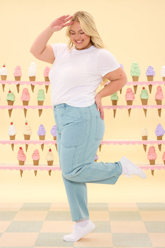 A smiling woman wearing the Ivy City Co Amanda Pants in Light Wash Denim and a white t-shirt poses playfully on one leg, saluting. She stands in front of pastel-colored ice cream cone decorations on a yellow background.