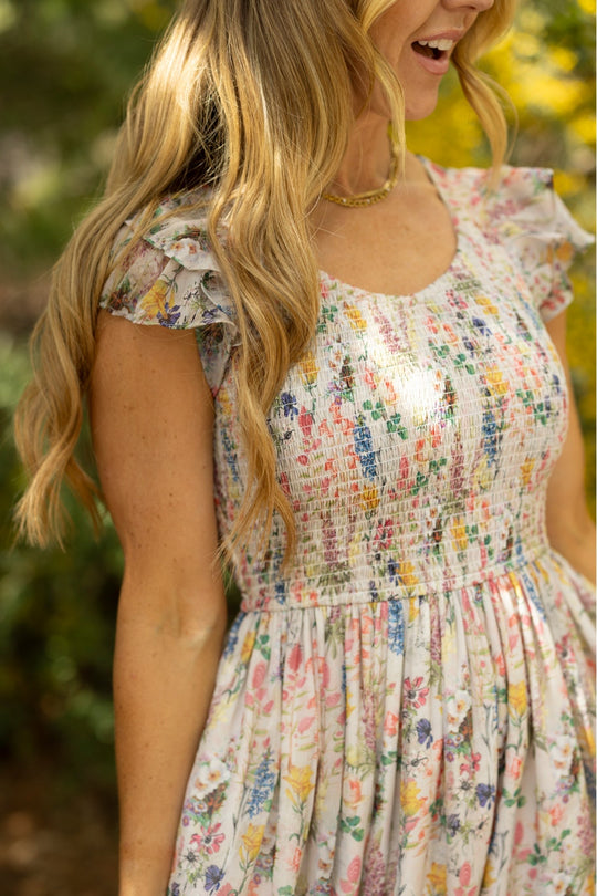 A woman with long, wavy blonde hair stands outdoors, smiling amid blurred green foliage and yellow flowers in the background. She wears the Ivy City Co Hattie Dress in Cordelia Floral, a maternity-friendly A-line midi dress with ruffled sleeves that combines comfort and effortless style for any occasion.