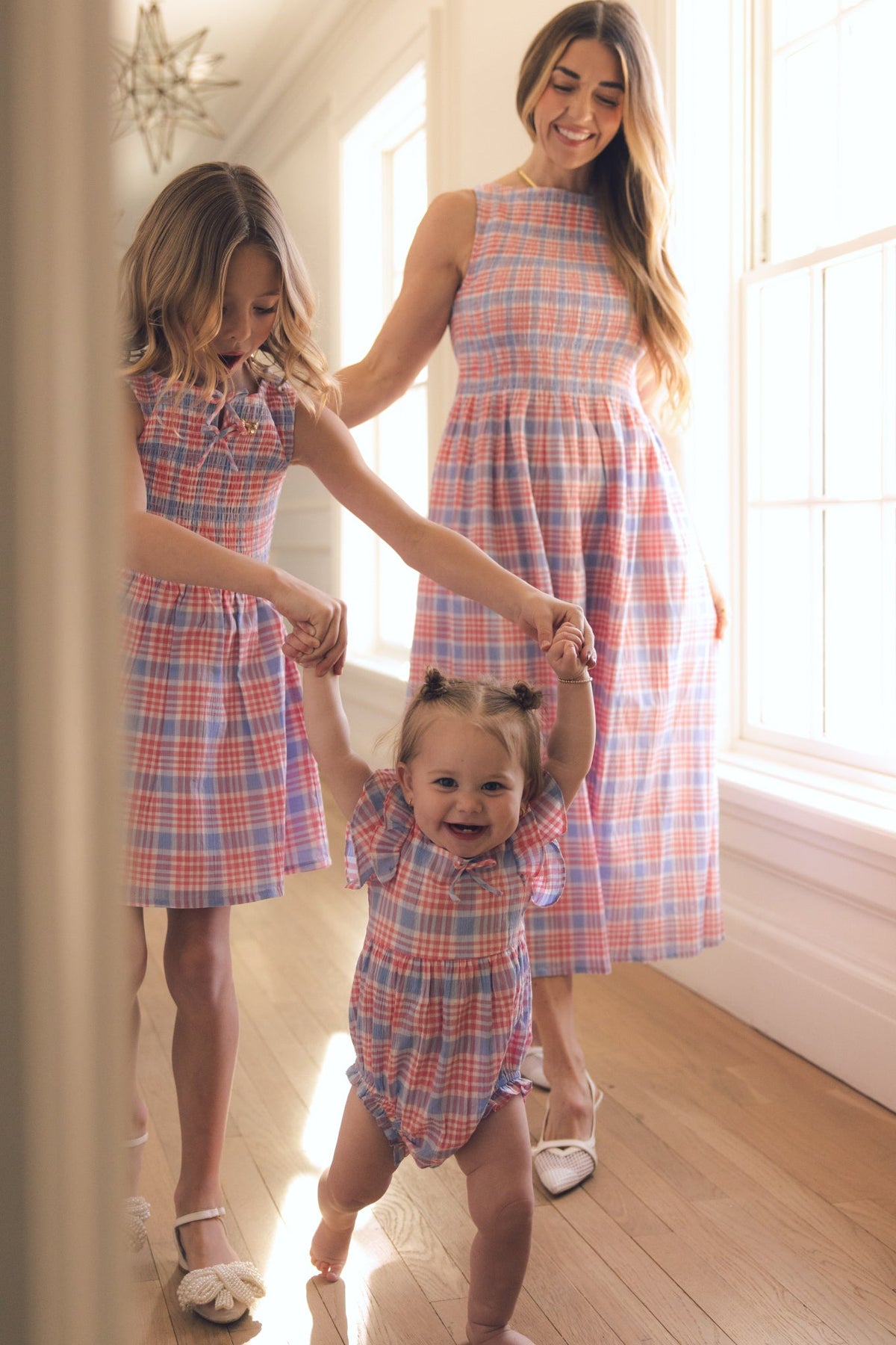 A woman, a young girl, and a toddler all wear matching Blossom Plaid outfits by Ivy City Co. The woman and girl hold the toddler’s hands as she smiles and walks in her Baby Ana Romper in Blossom Plaid, sunlight streaming through large windows beside them.