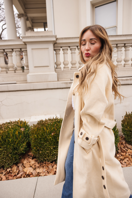A woman with long, wavy hair wears the Ivy City Co York Coat in Cream and blue jeans, standing outside a building with white columns and balustrades, eyes closed and lips pursed as if blowing a kiss.