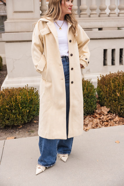 A woman stands outdoors wearing the Ivy City Co York Coat in Cream over a white t-shirt and blue jeans, styled with pointed cream shoes and a gold necklace. Bushes and a stone railing are in the background.