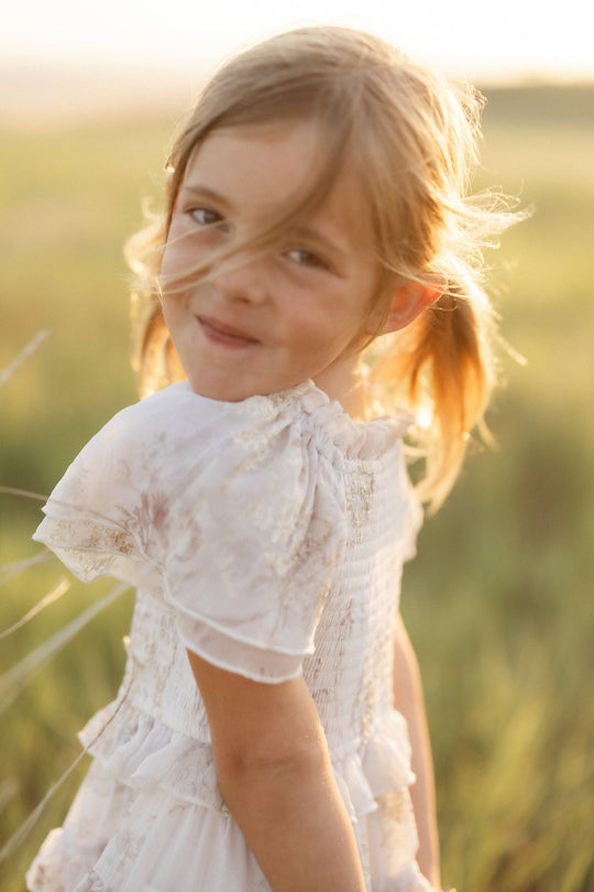 A young girl with blonde hair in pigtails smiles softly while standing outdoors in a sunlit field, wearing the Mini Winnie Dress in Bright White Wispy Wildflower by Ivy City Co.