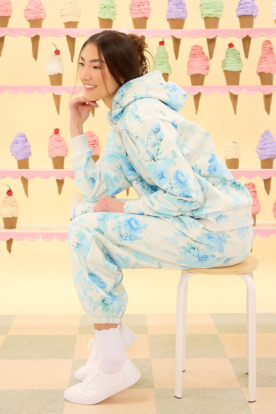 A person wearing the Ivy City Co Kian Jogger Sweatpants in Blue Aquarelle Fleur sits on a stool, smiling, in front of a pastel backdrop decorated with rows of colorful ice cream cone illustrations.