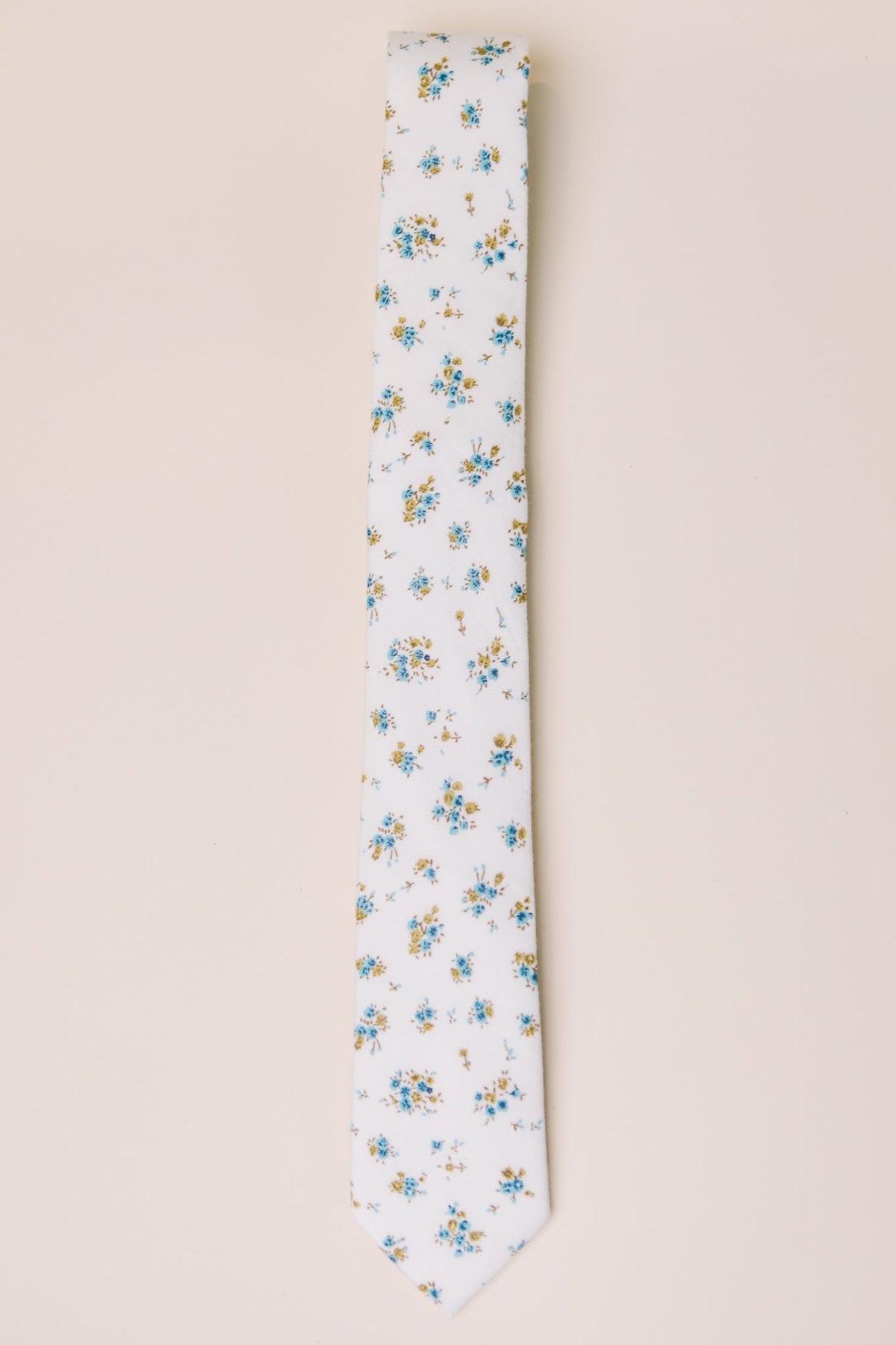 A white necktie featuring a delicate pattern of small blue and yellow flowers is laid flat on a light-colored background, perfectly complementing the Ivy City Co Ties in Madeline Blue Floral and making it an ideal choice for family matching accessories.