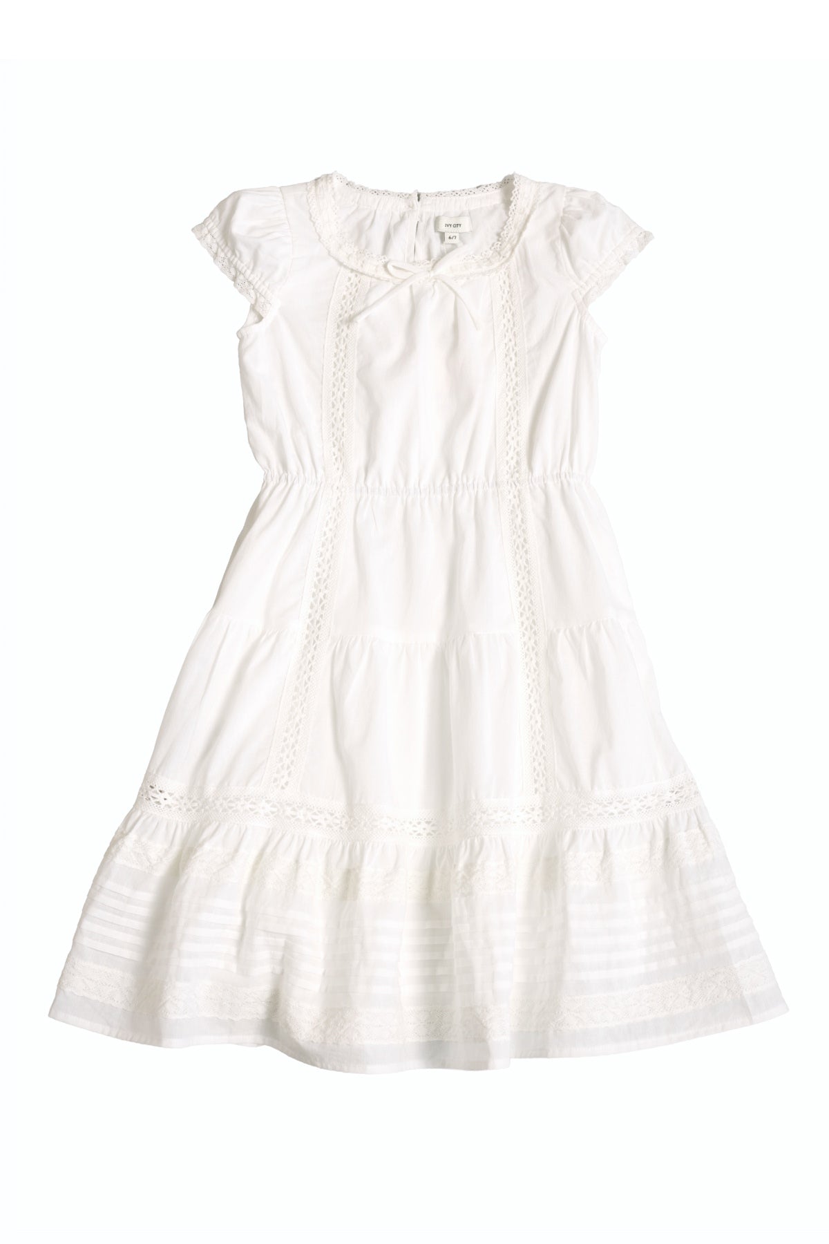 The Mini Palermo Dress in White by Ivy City Co features short sleeves, a knee-length cotton silhouette with lace details, a smocked bodice, round neckline, and ruffled hem, shown laid flat on a white background.