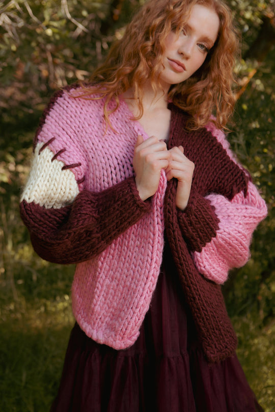 A woman with wavy red hair stands outdoors amidst greenery, wearing the Ivy City Co Marissa Sweater in Pink & Red.