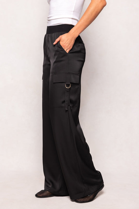 A person models the Ivy City Co Shay Satin Pants in Black—crafted with an elastic waistband, side pockets, and a loose fit—paired with a white sleeveless top while standing against a light background, one hand casually tucked into a pocket.