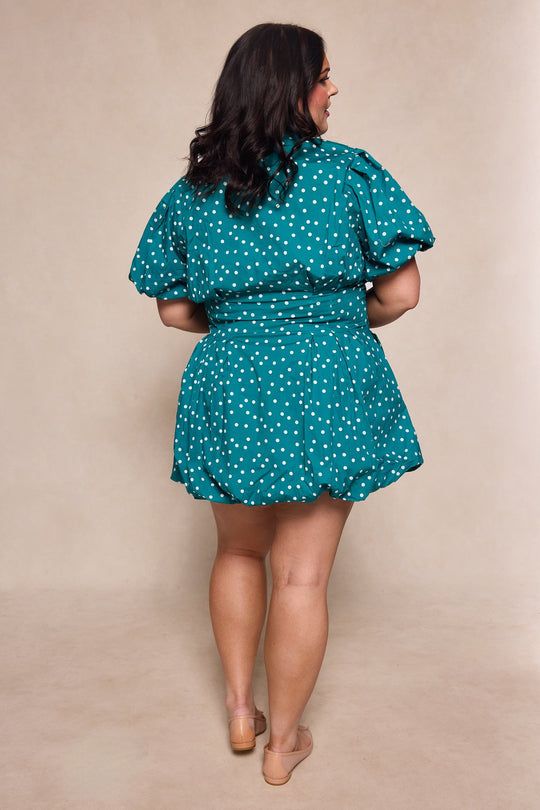 A woman with dark hair stands facing away, wearing the Ivy City Co Cody Dress in Deep Teal Polka Dot, featuring a short silhouette with puffed sleeves and nude shoes. The background is plain beige.