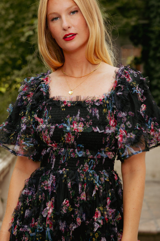 A woman with long blonde hair stands outdoors in soft natural light, wearing the Ivy City Co Florentina Dress in Obsidian Garden—a black tulle floral dress featuring ruffle details and puff sleeves—paired with a gold necklace.