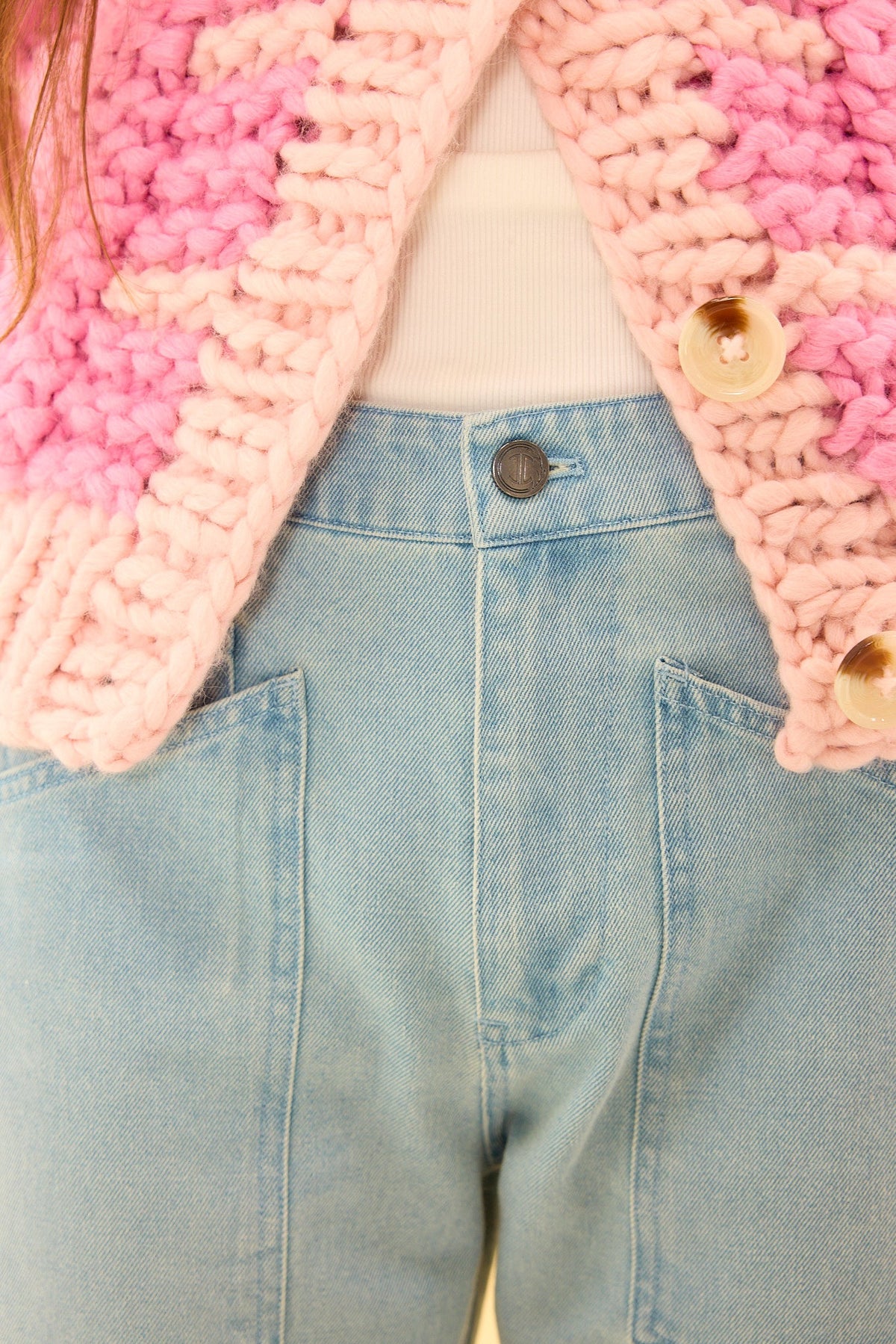 Close-up of a person wearing the Amanda Pants in Light Wash Denim by Ivy City Co, styled with a white ribbed top and a chunky pink knit sweater featuring large buttons. The image highlights the waist and upper body.
