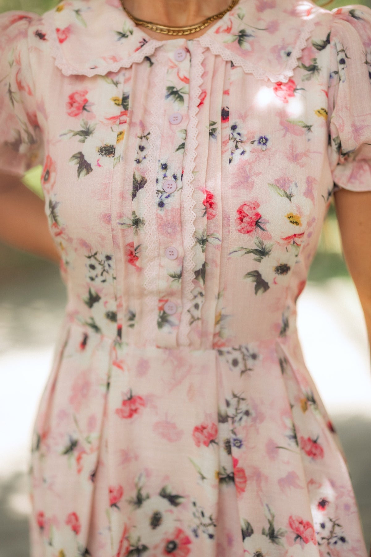 A person wearing the Ivy City Co Sophia Dress in Roselyn Pink—a light pink floral cotton-linen dress with pleats, buttons, and lace trim on the bodice, featuring a scalloped collar and puff sleeves.