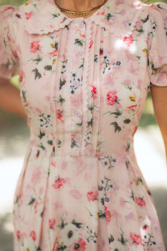 A person wearing the Ivy City Co Sophia Dress in Roselyn Pink—a light pink floral cotton-linen dress with pleats, buttons, and lace trim on the bodice, featuring a scalloped collar and puff sleeves.