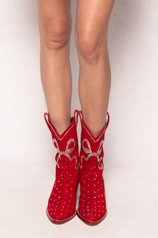 A person stands on a white background wearing the Ivy City Co Channing Bootie in Red—western-inspired ankle boots adorned with rhinestones and silver rope details. Only the legs and these bold, statement booties are visible.