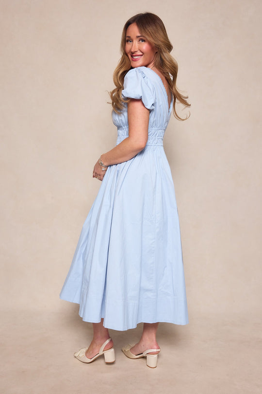 A woman with long hair, wearing the Ivy City Co Zella Dress in Sky Blue and cream heeled sandals, stands sideways and smiles at the camera in front of a beige background.