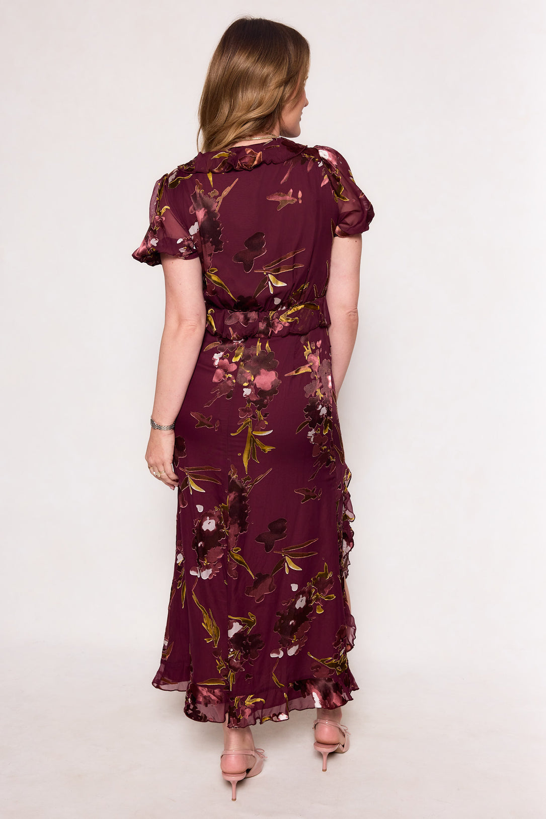 A woman faces away, wearing the Ivy City Co Zamora Dress in Burgundy Floral, featuring a fitted bodice and ruffled hem. Her light brown hair and pink heels complement this elegant celebration midi dress.