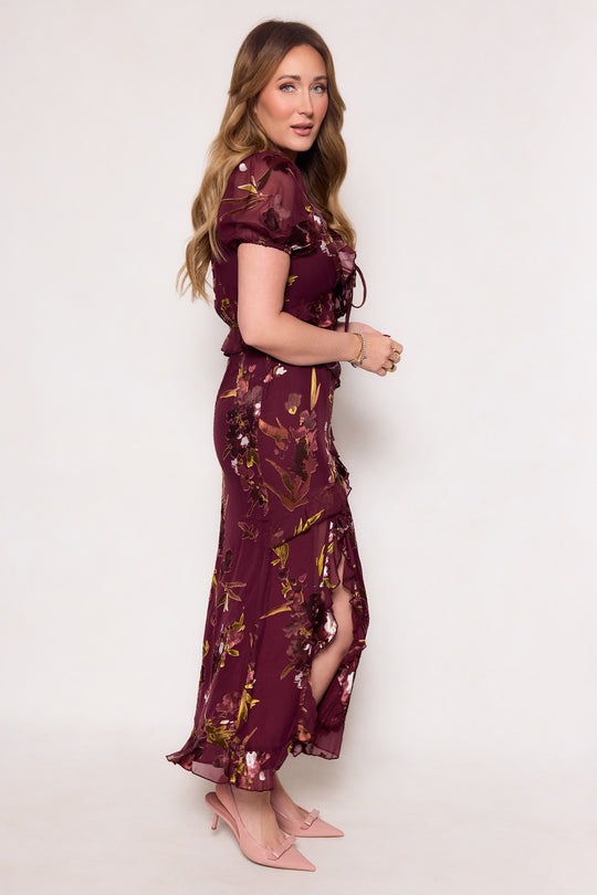 A woman with long, wavy hair stands sideways in the Zamora Dress in Burgundy Floral by Ivy City Co, featuring a fitted bodice and short sleeves, paired with pink pointed-toe heels against a plain light background.