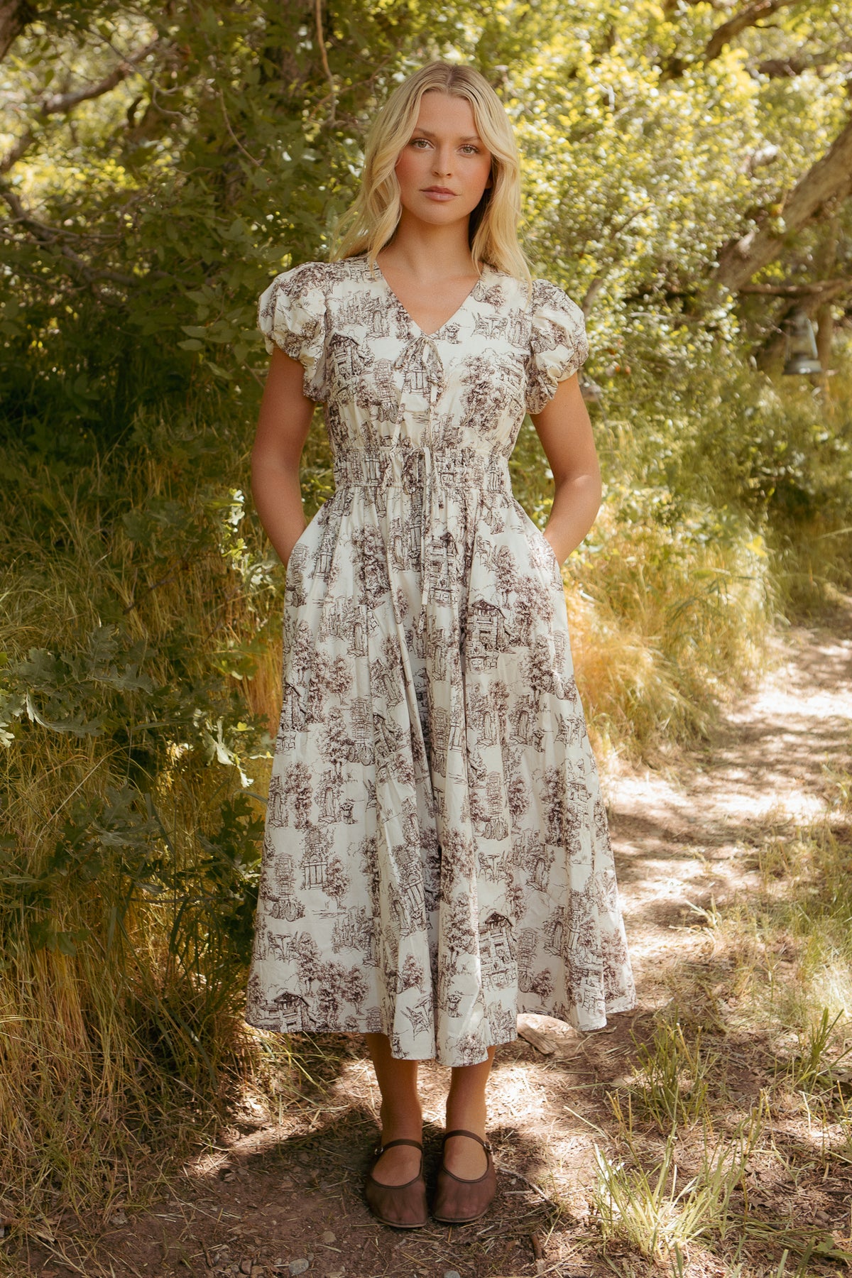 A woman stands on a sunlit dirt path surrounded by tall grass and trees, wearing the Ivy City Co Zaza Midi Dress in Brown Toile with pockets and delicate patterns, paired with brown Mary Jane shoes.